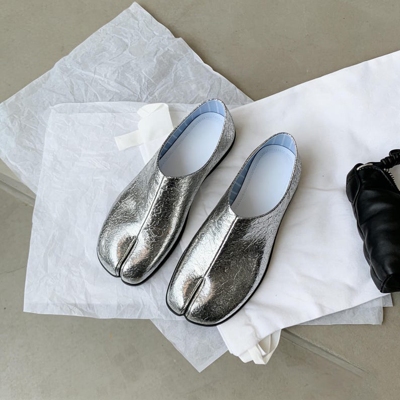 Puede incluir: Un par de bailarinas plateadas con puntera dividida y acabado texturizado. Los zapatos tienen interior blanco y suela negra. Los zapatos se exhiben sobre una superficie blanca.