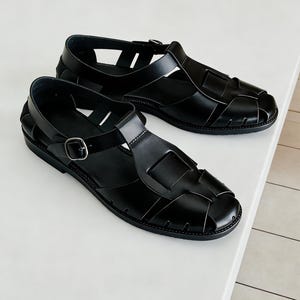 May include: A pair of black leather sandals with a woven design. The sandals have a rounded toe, a strap that buckles around the ankle, and a flat sole. The sandals are displayed on a white surface.