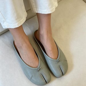 May include: A pair of light blue leather slip-on shoes with a split toe design. Each shoe features a small bow detail at the top. The shoes are shown on a white surface.