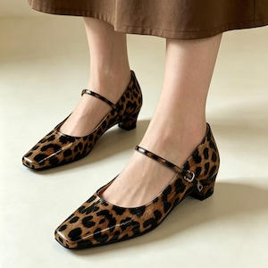 Puede incluir: Un par de zapatos Mary Jane con estampado de leopardo. Los zapatos tienen una puntera cuadrada, un tacón de bloque pequeño y una correa que se abrocha en la parte superior del pie. Los zapatos son marrones y negros.
