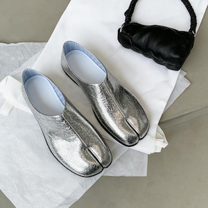 Op de afbeelding: Een paar zilverkleurige metallic schoenen met split-teen, witte binnenzolen en een zwarte handtas met een gedraaide handgreep. De schoenen hebben een uniek ontwerp.