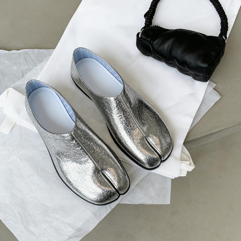 Puede incluir: Un par de zapatos plateados met&aacute;licos con punta dividida, plantillas blancas y un bolso negro con asa retorcida. Los zapatos tienen un dise&ntilde;o &uacute;nico, con una secci&oacute;n separada para el dedo gordo.