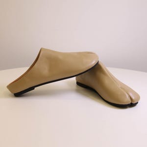 May include: A pair of beige leather slip-on shoes with a split-toe design. The shoes have a low heel and a black sole. The shoes are made of a smooth, matte leather material.