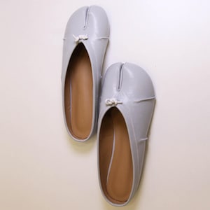 May include: A pair of light gray leather ballet flats with a split toe design. Each shoe features a small white knotted detail at the top. The interior of the shoes is a light brown color.