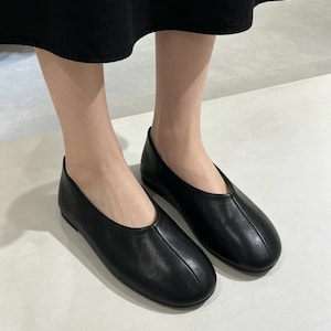 May include: A pair of black leather ballet flats. The shoes have a rounded toe and a low heel. The flats are smooth and sleek, with a simple design. The shoes are worn by a person.