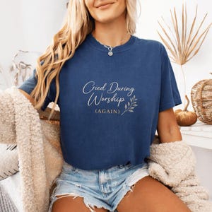 Puede incluir: Camiseta azul marino con el texto "Cried During Worship (AGAIN)" en fuente cursiva, acompañada de un diseño de hojas. La camiseta se combina con pantalones cortos de mezclilla.