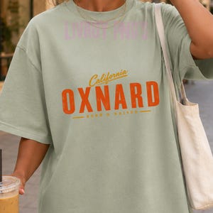 May include: A sage green short-sleeve t-shirt featuring the text California in yellow script above the word OXNARD in large orange block letters, with the text BORN & RAISED in yellow below, all centered on the front of the garment.
