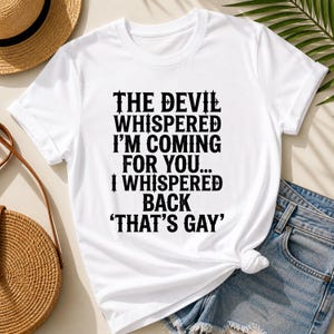 May include: White t-shirt featuring the text "THE DEVIL WHISPERED I'M COMING FOR YOU... I WHISPERED BACK 'THAT'S GAY'" in black. The shirt is paired with denim shorts and accessories.