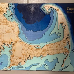 May include: A layered wooden bathymetric map of Cape Cod, Massachusetts, USA. The map features light wood landmasses with engraved roads and text, set against deep blue, navy, and light blue water layers. A compass rose is in the top left corner.