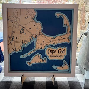 May include: A framed wooden map of Cape Cod, Massachusetts, with a blue background and light wood detailing. The map is set in a white frame and includes the text "Cape Cod" and "Massachusetts". The map is displayed on a black and white checkered surface.