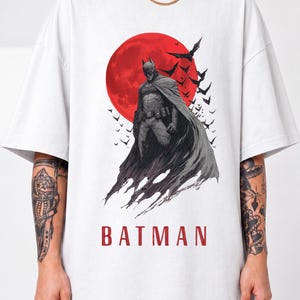 Peut inclure: Un t-shirt blanc avec une impression graphique de Batman debout devant une grande lune rouge. Le motif comprend un essaim de chauves-souris volant vers la droite et le mot BATMAN imprimé en lettres capitales rouges au bas de l'illustration.