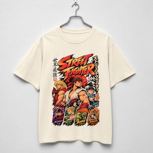 Può includere: Una t-shirt a maniche corte color crema con una grande stampa grafica dei personaggi di Street Fighter, tra cui Ryu, Ken e Chun-Li. Il design include testo in rosso e giallo, caratteri giapponesi e un elemento a forma di sole rosso.