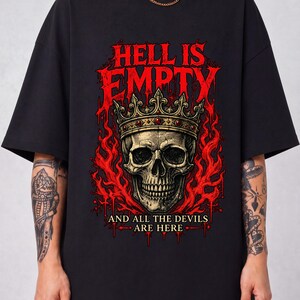 Puede incluir: Una camiseta negra con un estampado gráfico de una calavera con corona, rodeada de llamas rojas. El texto HELL IS EMPTY está impreso en rojo sobre la calavera, y el texto AND ALL THE DEVILS ARE HERE está impreso en blanco debajo.