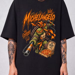 May include: A black t-shirt featuring a graphic print of the Teenage Mutant Ninja Turtle Michelangelo. The design includes the text Michelangelo, Cowabunga, and Pizza, with the character holding nunchucks next to an open box of pepperoni pizza.