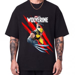 May include: A black short-sleeved t-shirt featuring a graphic of the Marvel Studios Wolverine character in a yellow and blue suit with extended claws. Behind the character are red claw marks and the white text Wolverine above the character.
