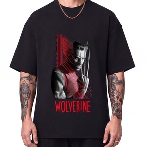 May include: A black short-sleeved t-shirt featuring a graphic print of the character Wolverine with metallic claws extended. The design incorporates red vertical lines and the text WOLVERINE in red lettering centred below the character illustration.