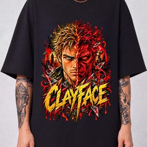 Puede incluir: Una camiseta negra de manga corta con un estampado gráfico de un personaje con el rostro dividido. Un lado muestra una cara humana con un ojo azul, mientras que el otro es una cara roja, texturizada y fundida. El texto CLAYFACE aparece en amarillo.