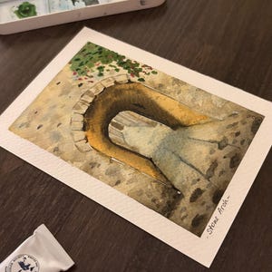 Może przedstawiać: Akwarela przedstawiająca kamienny łuk w ciepłych odcieniach. Dzieło przedstawia szczegółowe odwzorowanie łuku, z widocznymi pociągnięciami pędzla i fakturami. Obraz jest na białym papierze, z tubkami farb akwarelowych i paletą w tle.