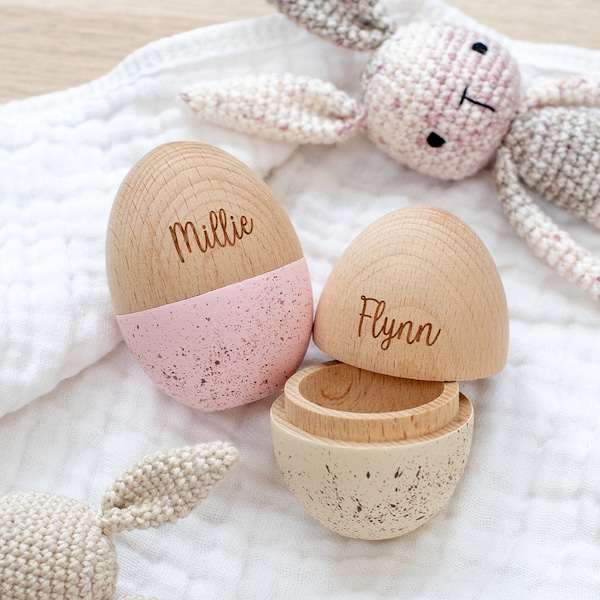 May include: Two wooden egg-shaped boxes with lids, one painted pink with white speckles and the other beige with brown speckles. The boxes are engraved with the names "Millie" and "Flynn".