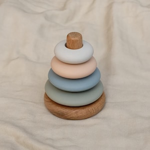 May include: A wooden stacking toy with five rings in shades of blue, green, and pink. The rings are made of silicone and stack on a wooden base.