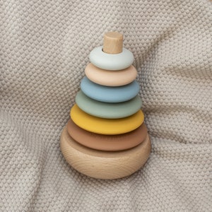 May include: A wooden stacking toy with six colorful rings. The rings are in shades of blue, green, yellow, brown, and beige. The toy is on a light gray textured surface.
