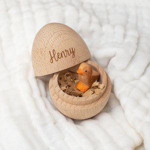 May include: A wooden egg-shaped box with the name "Henry" engraved on the top. The box is open and a small wooden bird is inside, sitting on a bed of shredded paper.