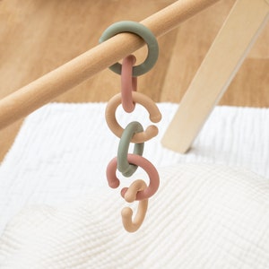 May include: A baby toy featuring a chain of colorful, interlocking rings in shades of green, pink, and tan. The rings are suspended from a wooden dowel. The toy is designed for infants and toddlers.