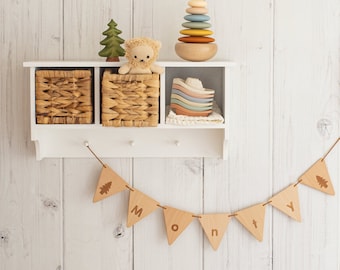 May include: A white shelf with three compartments, two of which contain woven baskets. The shelf also has a wooden toy stacking ring and a set of nesting bowls. A wooden banner with the word "MONTY" hangs below the shelf.