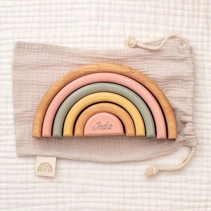 Könnte beinhalten: Ein Holz-Regenbogen-Stapelspielzeug mit fünf Bögen in den Farben Rosa, Grün und Gelb. Die Bögen sind ineinander verschachtelt und der kleinste Bogen trägt den Namen "India" eingraviert. Der Regenbogen liegt auf einer hellbraunen Stoffoberfläche.