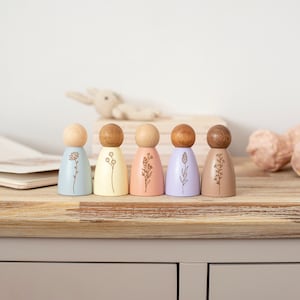 Puede incluir: Seis figuras de muñecas de madera con diferentes diseños florales pintados en ellas. Las muñecas están pintadas en colores pastel, incluyendo azul claro, amarillo, rosa, morado y marrón.