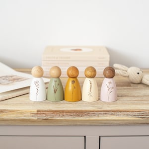Puede incluir: Cinco muñecas de madera con diferentes diseños florales pintados en ellas. Las muñecas están en fila sobre una superficie de madera. Las muñecas están pintadas de blanco, verde, amarillo, crema y rosa.