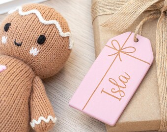 May include: A pink wooden gift tag with the name Isla written in cursive on it. The tag is tied with a brown ribbon and is hanging from a brown gift box.