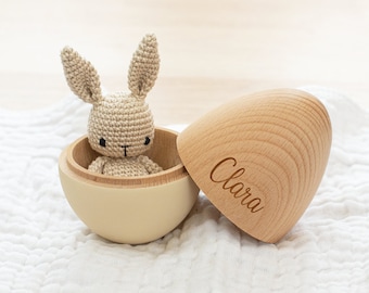 May include: A beige crocheted bunny sits inside a two-toned wooden egg. The egg is open, revealing the bunny. The egg's lid is engraved with the name "Clara". The egg is light beige on the outside and natural wood on the inside.