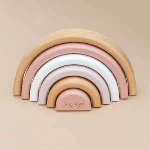 May include: A wooden rainbow stacking toy with five arches in shades of pink, white, and natural wood. The smallest arch has the name "Brayleigh" engraved on it.