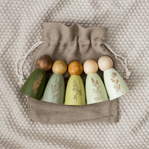 Puede incluir: Cinco figuras de muñecas de madera con cuerpos pintados de verde y diferentes diseños florales. Las muñecas están dispuestas en fila y están sentadas en una pequeña bolsa de arpillera.