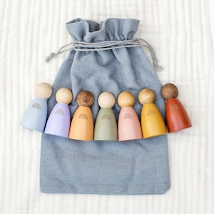 Könnte beinhalten: Ein Set aus acht Holzpuppen mit bemalten Regenbogenmustern. Die Puppen sind in einer Reihe angeordnet und in verschiedenen Farben, darunter Blau, Lila, Gelb, Grün, Rosa und Orange. Die Puppen sitzen in einer grauen Stofftasche.