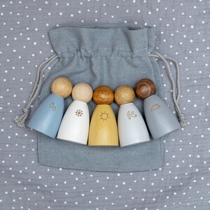 Könnte beinhalten: Satz von fünf Holzfiguren, die Wettermenschen darstellen, in einer grauen Stofftasche. Die Figuren sind in verschiedenen Farben bemalt und haben Wettersymbole darauf: ein Regentropfen, ein Schneeflocke, eine Sonne, ein Wind und eine Wolke.