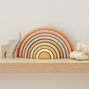 Pode incluir: Um brinquedo de empilhar arco-íris feito de madeira com cores pastel. O arco-íris é feito de sete arcos, cada um de uma cor diferente. O brinquedo está sentado em uma prateleira de madeira.