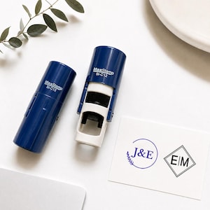 Puede incluir: Dos sellos autoentintables MaxStamp SI-C12 de color azul oscuro. Uno tiene tapa y el otro está abierto, mostrando una base blanca. Una tarjeta blanca muestra dos diseños estampados: un monograma circular azul con J&E y un monograma de diamante negro con E|M.