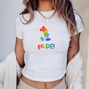 May include: White crop top featuring a rainbow-colored Mickey Mouse design and the word "PRIDE!" in rainbow letters. The design is a silhouette of Mickey Mouse. The shirt has short sleeves and a crew neck.