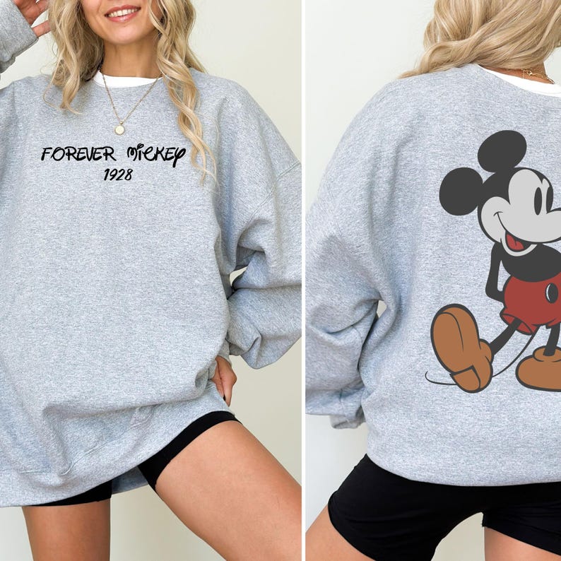 Puede incluir: Una sudadera gris con el texto "FOREVER MICKEY 1928" en la parte delantera. La parte trasera de la sudadera muestra un dibujo de Mickey Mouse. La sudadera tiene cuello redondo y mangas largas.
