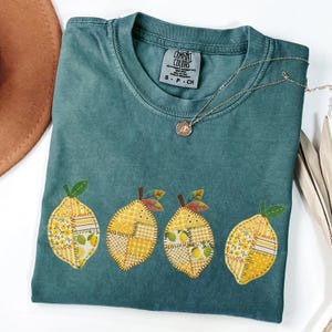 May include: A teal Comfort Colors t-shirt with four lemon appliqués. Each lemon is a patchwork of yellow fabrics with green leaves. A gold necklace with a small pendant rests on the shirt. The shirt is folded.