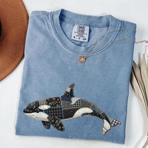 May include: A pale blue t-shirt with a patchwork orca design. The orca is made of various black and white patterned fabrics. A gold necklace with a pendant is draped across the shirt. The shirt has the Comfort Colors brand label.