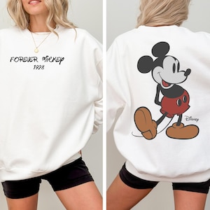 Puede incluir: Sudadera blanca con "FOREVER MICKEY 1928" en escritura negra en la parte delantera. La parte trasera muestra un gran gr&aacute;fico de Mickey Mouse en rojo, negro y marr&oacute;n, con el logotipo de Disney. Una prenda casual y c&oacute;moda.