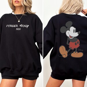 Puede incluir: Sudadera negra con ribete blanco. La parte delantera tiene el texto "FOREVER MICKEY 1928". La parte trasera muestra un gr&aacute;fico de Mickey Mouse con pantalones cortos rojos y zapatos marrones. El logotipo de Disney es visible.
