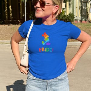 May include: A royal blue t-shirt featuring a rainbow-colored cartoon character and the word "PRIDE!" in rainbow colors. The shirt is paired with light blue denim jeans. The overall theme is LGBTQ+ pride.