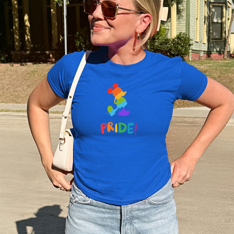 May include: A royal blue t-shirt featuring a rainbow-colored cartoon character and the word "PRIDE!" in rainbow colors. The shirt is paired with light blue denim jeans. The overall theme is LGBTQ+ pride.