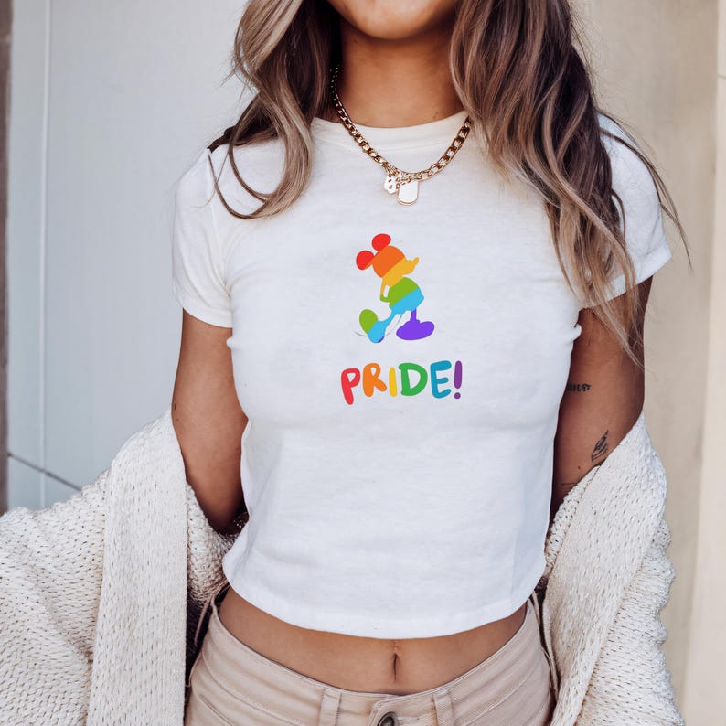 May include: White crop top featuring a rainbow-colored Mickey Mouse design and the word "PRIDE!" in rainbow letters. The design is a silhouette of Mickey Mouse. The shirt has short sleeves and a crew neck.