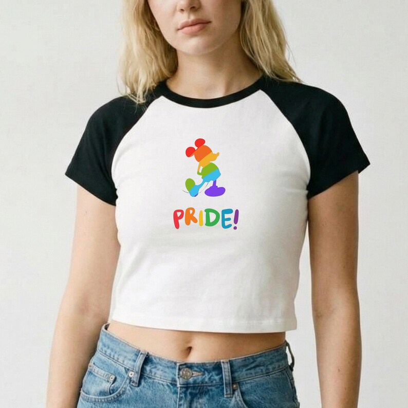 May include: White crop top with black raglan sleeves featuring a colorful Mickey Mouse design in rainbow colors. The word "PRIDE!" is printed below the character. The shirt is paired with blue jeans.