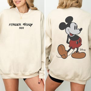 Puede incluir: Sudadera color crema con el texto "FOREVER MICKEY 1928" en escritura negra en la parte delantera. La parte trasera muestra un gran gr&aacute;fico de Mickey Mouse en su pose cl&aacute;sica, con el logo de Disney.